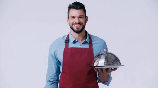 Camarero sonriente en delantal sosteniendo el plato de servir con cloche aislado en gris - foto de stock