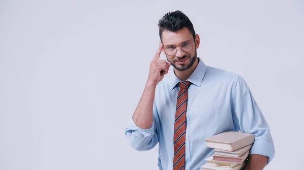 Profesor sonriente sosteniendo un montón de libros y apuntando a la cabeza aislado en gris - foto de stock