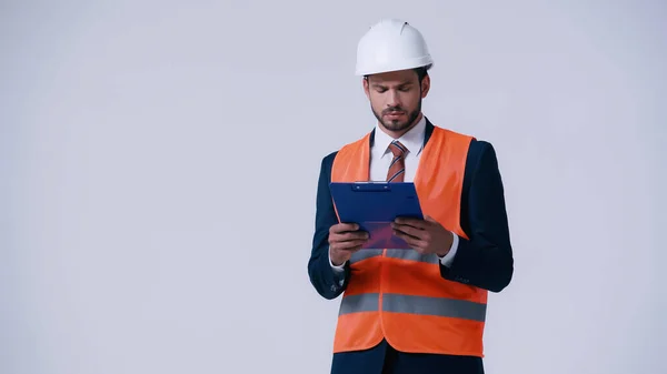 Ingénieur réfléchi dans le casque et gilet de sécurité en regardant le presse-papiers isolé sur gris — Photo de stock