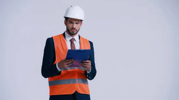 Contratista serio en hardhat sujetando portapapeles y mirando hacia otro lado aislado en gris - foto de stock