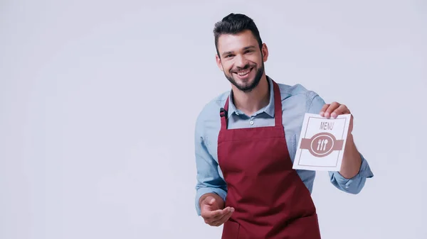 Glücklicher junger Kellner in Schürze zeigt Speisekarte isoliert auf grau — Stockfoto