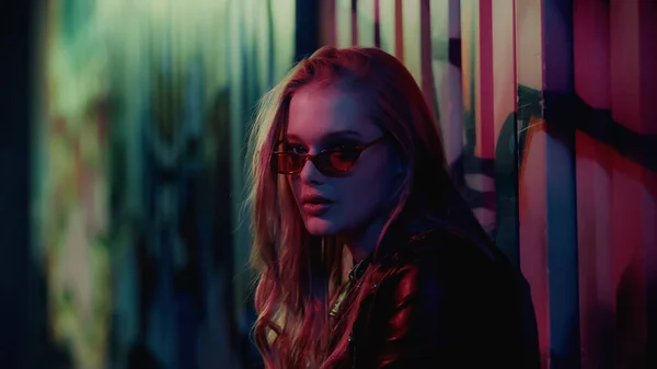 Молода жінка в сонцезахисних окулярах дивиться на камеру біля стіни з графіті під час вечірки — стокове фото