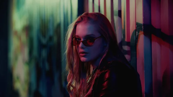 Young woman in sunglasses looking at camera near wall with graffiti during party — Stock Photo