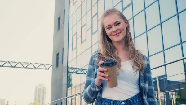 Vue à angle bas de la femme blonde souriante tenant du café pour aller et regardant la caméra sur la rue urbaine — Photo de stock