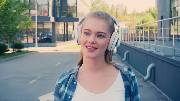 Femme souriante écoutant de la musique dans des écouteurs sans fil dans la rue urbaine — Photo de stock