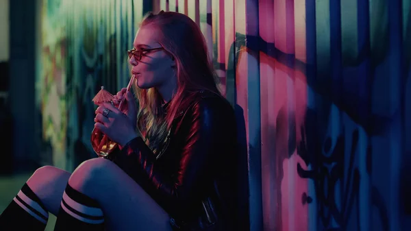 Junge Frau mit Sonnenbrille trinkt während Party Cocktail an Wand mit Graffiti — Stockfoto