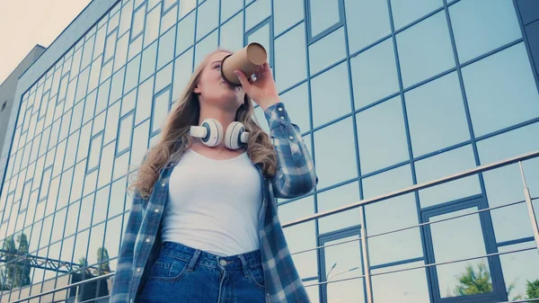 Visão de baixo ângulo da jovem com fones de ouvido bebendo café para ir na rua urbana — Fotografia de Stock