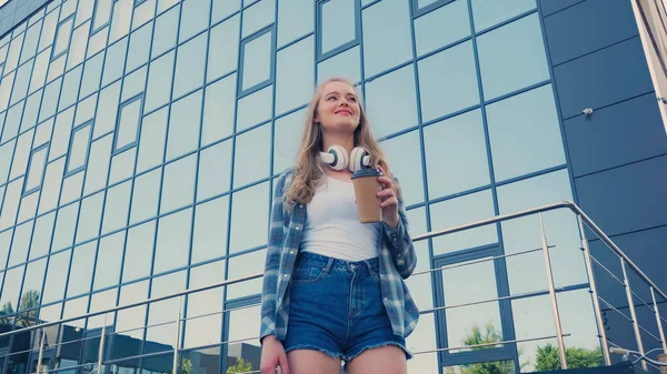 Vue à angle bas de la femme positive avec écouteurs tenant café pour aller près du bâtiment à l'extérieur — Photo de stock