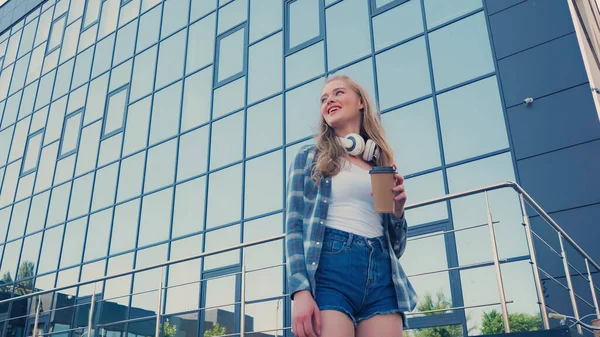 Low angle view of happy blonde woman with headphones holding paper cup near building outdoors — Stock Photo