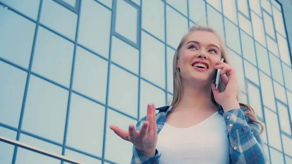 Vista de ángulo bajo de la mujer rubia sonriente haciendo gestos y hablando en el teléfono inteligente al aire libre - foto de stock