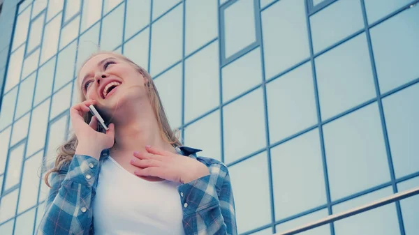 Low angle view of positive woman in plaid shirt talking on cellphone on urban street — Stock Photo