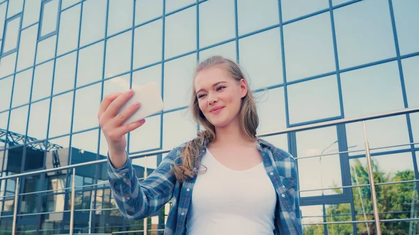 Позитивная молодая женщина в клетчатой рубашке делает селфи на смартфоне на городской улице — стоковое фото