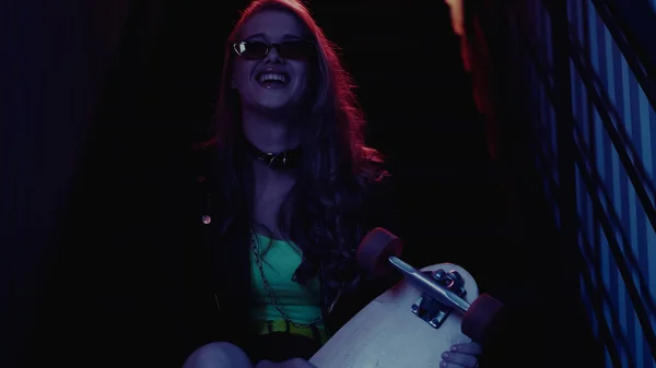 Cheerful woman in sunglasses holding longboard in nightclub — Stock Photo