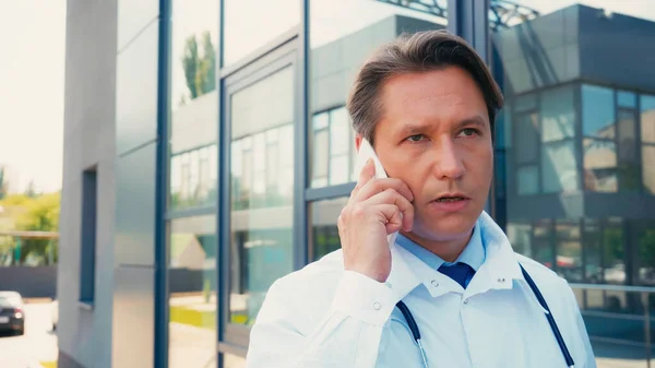 Dottore serio in soprabito bianco che parla su smartphone vicino a costruzione di clinica — Foto stock