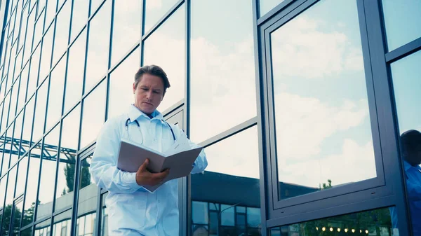 Doctor looking at documents in folder near hospital building — Stock Photo