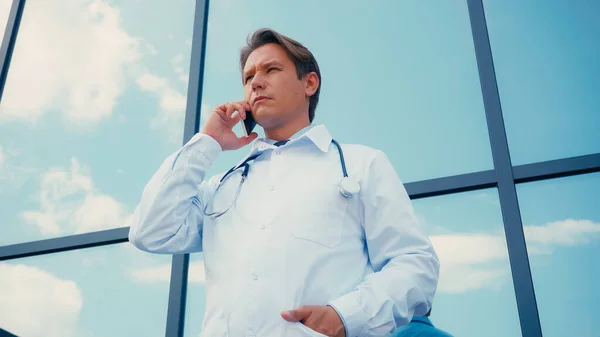 Vista de ángulo bajo del médico con la mano en el bolsillo de la bata blanca hablando en el teléfono celular al aire libre - foto de stock