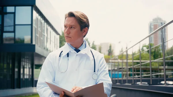 Doctor en bata blanca con estetoscopio y carpeta mirando al aire libre - foto de stock