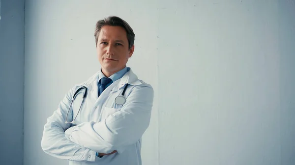 Уверенный в себе доктор в белом халате, стоящий со скрещенными руками возле серой стены — стоковое фото