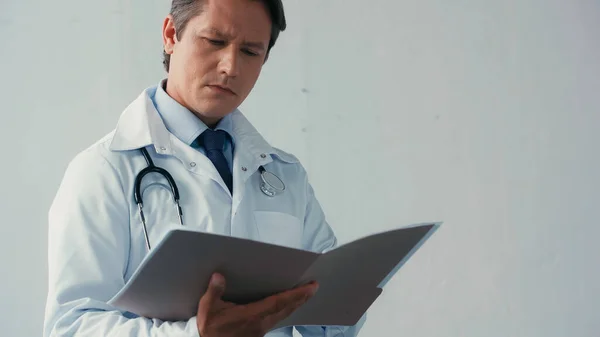 Médico pensativo de bata blanca leyendo documentos en carpeta sobre fondo gris - foto de stock