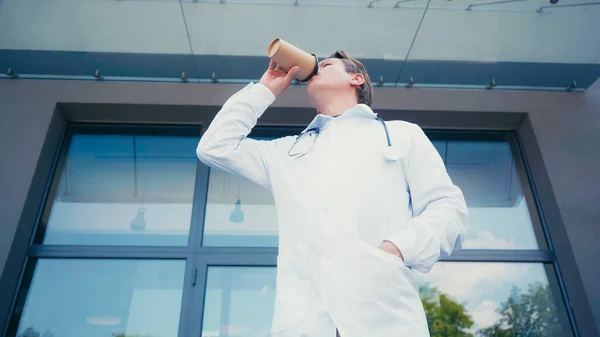 Vista a basso angolo del medico con mano in tasca di cappotto bianco bere caffè per uscire all'aperto — Foto stock