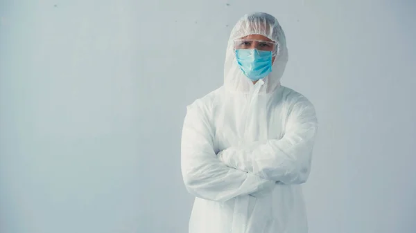 Doctor in hazmat suit and medical mask with goggles standing with crossed arms on grey background — Stock Photo