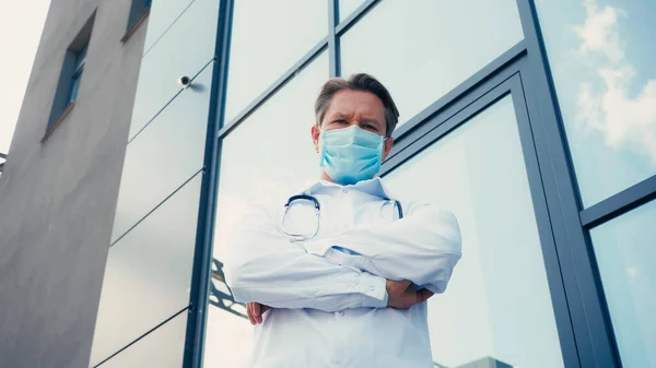 Low angle view of doctor in medical mask standing with crossed arms and looking at camera — Stock Photo