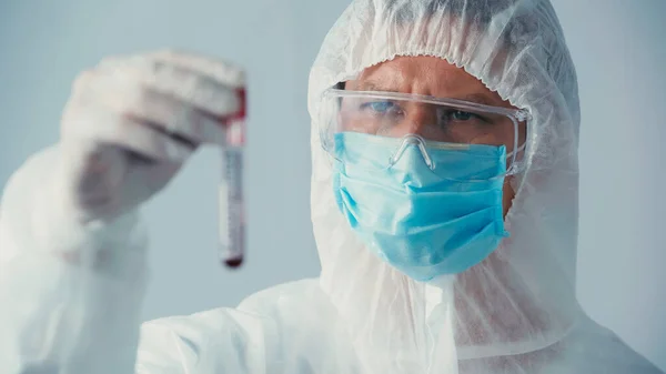 Virologist in hazmat suit and goggles looking at blurred test tube isolated on grey — Stock Photo