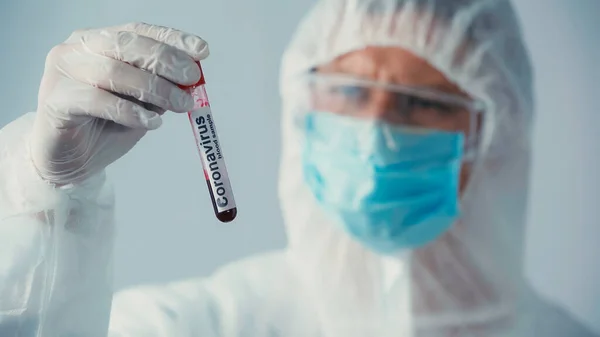 Селективный фокус коронавирусной пробирки в руке врача в латексной перчатке и защитном костюме, изолированном на сером — стоковое фото