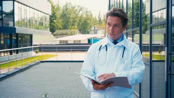 Médico en el abrigo blanco que sostiene la carpeta con los documentos y mirando hacia fuera al aire libre - foto de stock