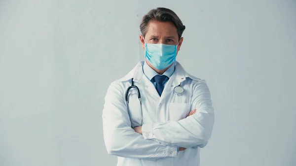 Doctor in medical mask and white coat standing with crossed arms on grey — Stock Photo