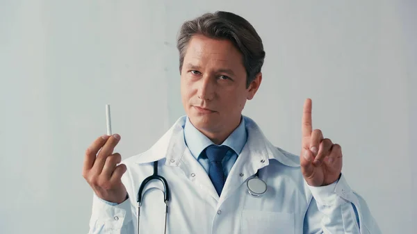 Doctor holding cigarette and showing warning gesture while looking at camera on grey — Stock Photo