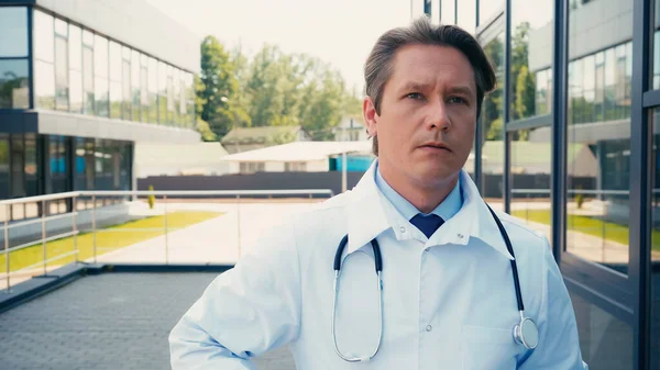 Doctor in white coat and with stethoscope looking away near city hospital — Stock Photo