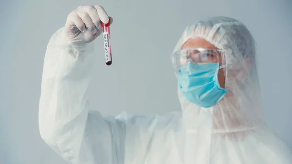 Científico borroso en traje de felpudo y guante de látex mirando tubo con prueba de coronavirus aislado en gris - foto de stock