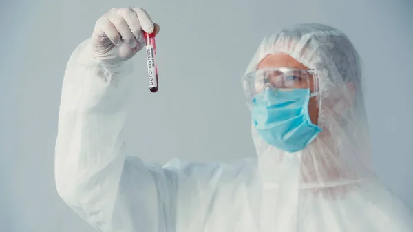 Blurred scientist in hazmat suit and latex glove looking at tube with coronavirus test isolated on grey — Stock Photo