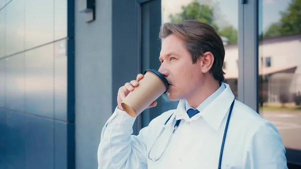 Doctor in white coat drinking coffee near building of city clinic — Stock Photo