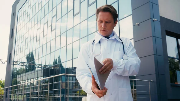 Médecin en manteau blanc regardant le dossier avec des documents à l'extérieur — Photo de stock