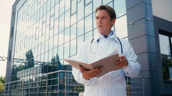 Doctor in white coat holding folder and looking away near building of hospital — Stock Photo