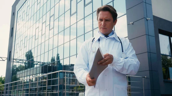 Серьезный врач в белом халате, держащий папку с документами возле больницы со стеклянным фасадом — стоковое фото