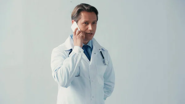 Внимательный доктор в белом халате разговаривает по смартфону на сером фоне — стоковое фото