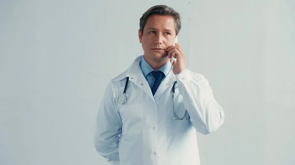 Doctor talking on mobile phone while standing with hand behind back on grey — Stock Photo