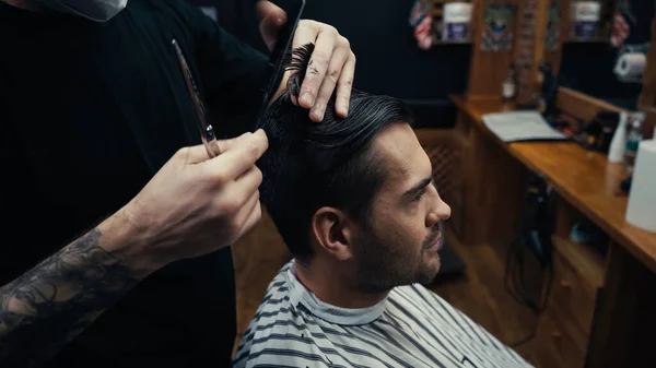 Tattooed hairdresser combing hair of client in barbershop — Stock Photo