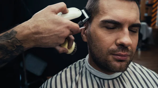 Tattooed barber trimming hair of man in barbershop — Stock Photo