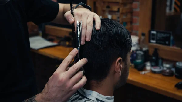 Barber holding comb and cutting hair of man in blurred barbershop — Stock Photo