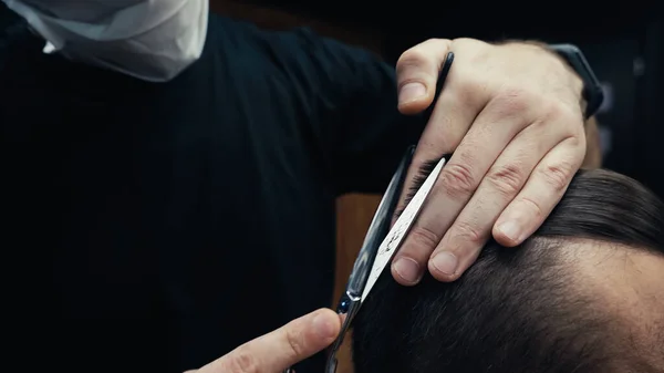 Cropped view of barber in medical mask cutting hair of man in barbershop — Stock Photo