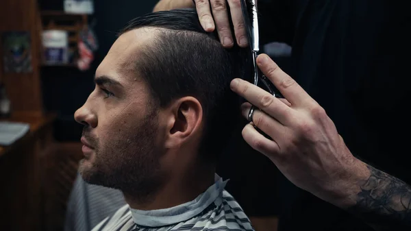 Hairdresser cutting hair of man in hairdressing cape in barbershop — Stock Photo