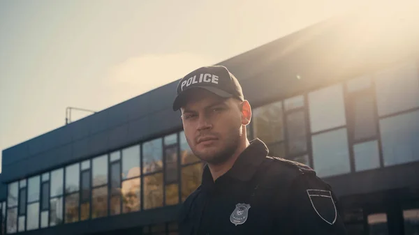 Joven policía con gorra con letras y uniforme mirando a la cámara cerca del edificio - foto de stock