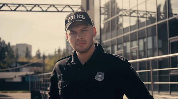 Jeune policier en casquette avec lettrage et uniforme regardant la caméra à l'extérieur — Photo de stock