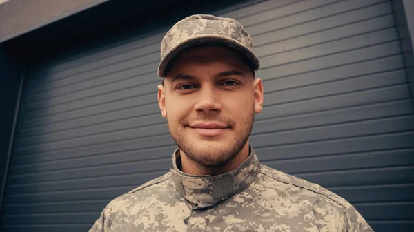 Soldat en uniforme et casquette souriant en regardant la caméra près du bâtiment — Photo de stock