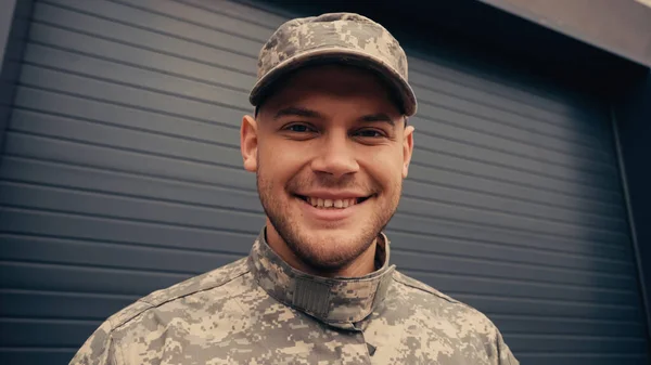 Cheerful soldier in uniform and cap looking at camera near building — Stock Photo