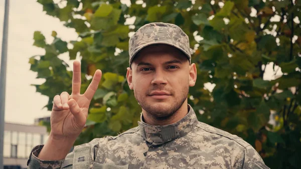 Young soldier in uniform and cap showing peace sign near tree outdoors — Stock Photo